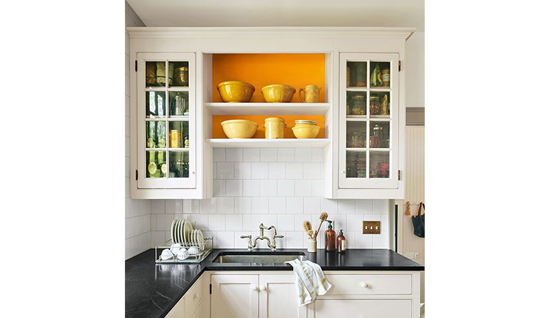 A mostly white-painted kitchen with black counter tops and orange accent behind shelving.