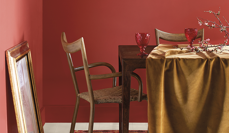 Comedor con pared roja clara, armarios, chimenea y luz dorada blancos, mantel blanco y flores morada