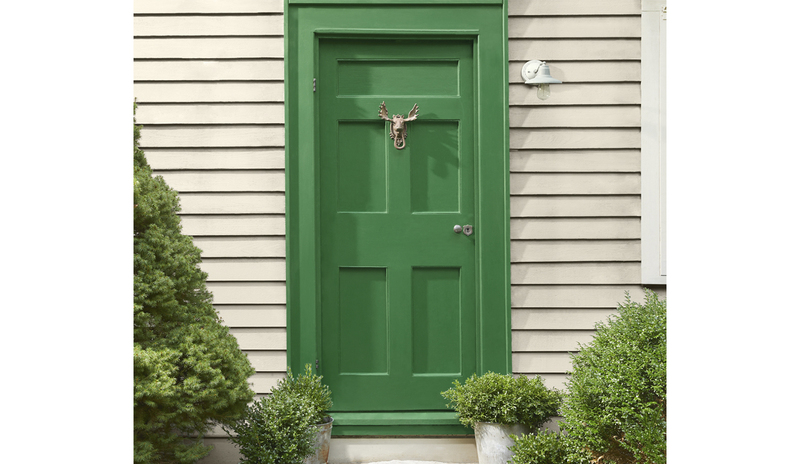 Warm greige-painted house siding, with a pretty green door flanked by shrubs, and white window trim.