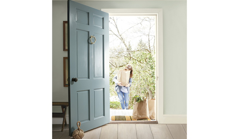 A soft blue-painted front door opens into a home as a woman carrying groceries walks towards it.