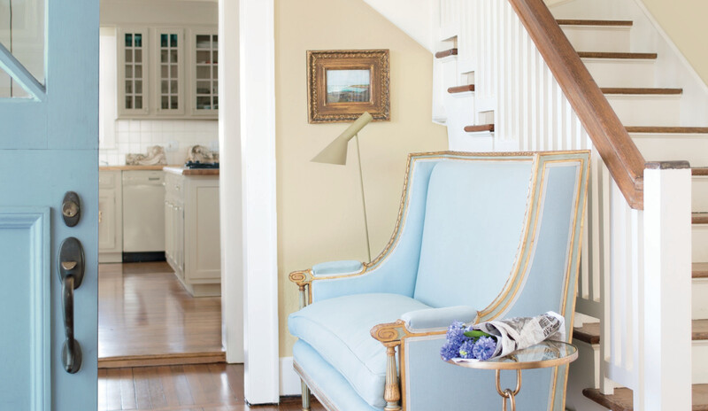 A bright entryway in neutral shades includes a staircase, blue chaise and light blue front door.