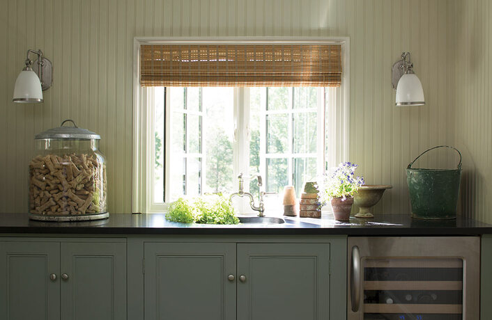 A kitchen counter in shades of light green features a sink window, sconce lights and plants.