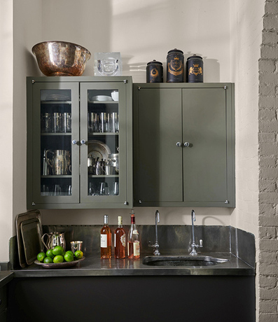 Una barra con fregadero con mostradores negros, gabinetes pintados de gris, vino y varios vasos.