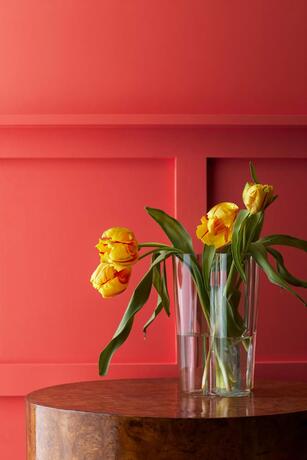 Flores amarillas en un jarrón sobre una mesa frente a paredes y paneles pintados de color frambuesa.