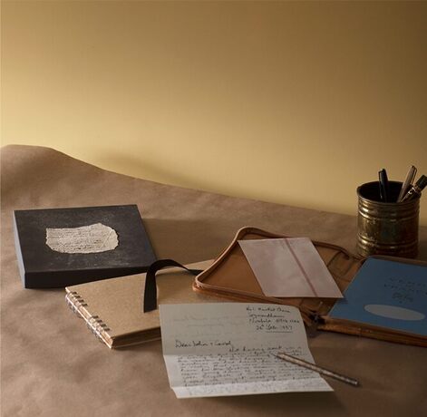 A collection of notebooks and pens atop brown paper, next to an earthy honey color painted wall.