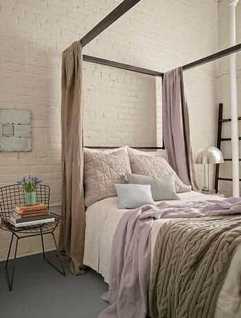 Dormitorio con pared de ladrillo blanqueado, cama con dosel beige y decoración lila.