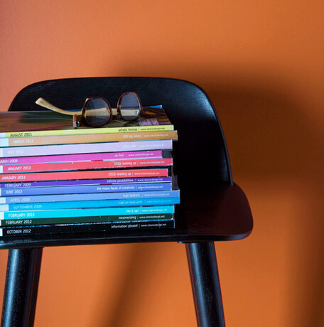 Una silla negra con una pila de revistas multicolores se destaca en una pared naranja.