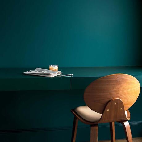 Pared pintada de verde azulado oscuro y escritorio con periódico y silla de madera clara.