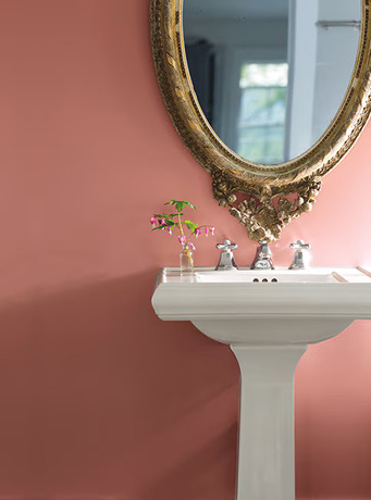 Un baño pintado de rosa suave con espejo dorado ornamentado y lavabo blanco.