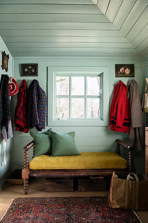 Vestíbulo pintado de verde con banco de madera, cojín amarillo de terciopelo y chaquetas colgadas.