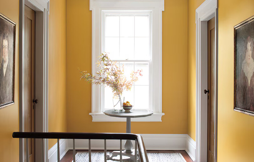 Yellow-painted hallway with wooden table and flowers, two doors, a window, and white-painted trim.