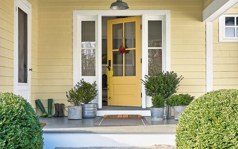 The front porch of a yellow-painted house with a yellow door, white trim, plants in metal pails, 