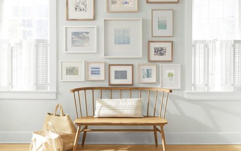 An off-white painted wall with a collection of framed art behind a wooden bench and shopping bags.